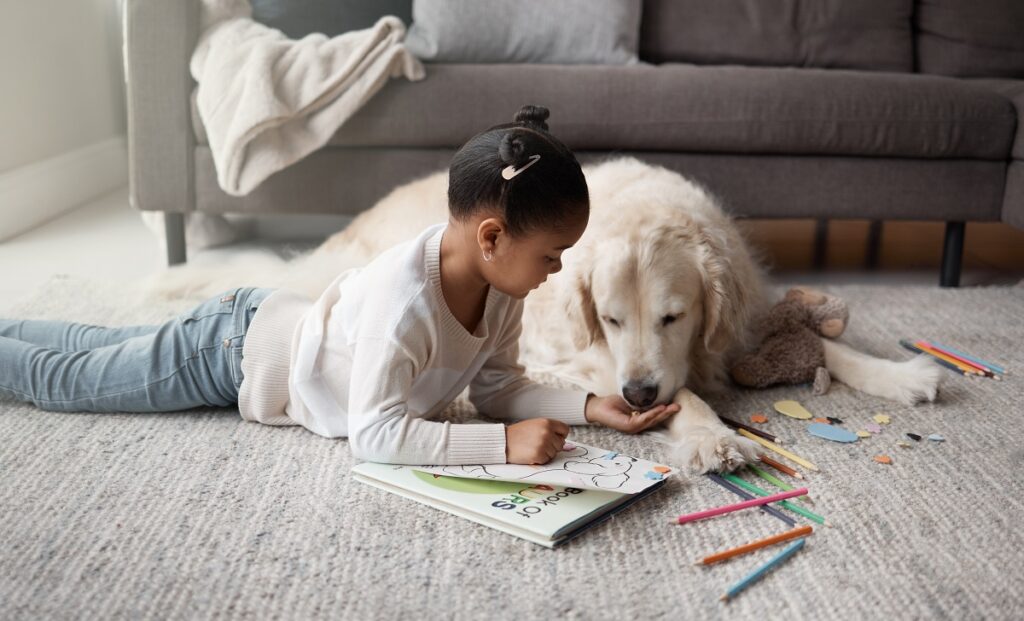 Una bambina distesa su un tappeto colora un disegno accanto al suo cane