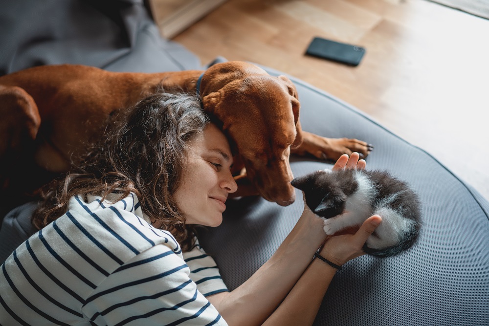Vivere con cani o gatti: una proprietaria tiene in mano un piccolo gattino distesa in salotto con accanto il suo cane