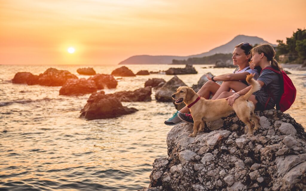 Una mamma con la figlia e il loro cane guardano  il tramonto al mare