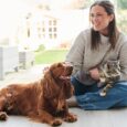 Una donna sorride coccolando il suo cane e il suo gatto: per il loro benessere è importante fare attenzione anche alle allergie alimentari nei pet